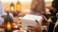 Hands holding white gift box during festive celebration with lanterns and people in background at golden hour, suitable for holiday marketing, greeting cards, and seasonal promotions.