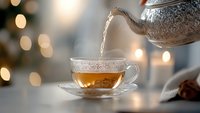 Pouring hot aromatic tea from vintage silver teapot into transparent glass cup on blurred bokeh background with soft warm lights and candles.