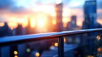 Modern glass railing with metal handrail on urban balcony or terrace overlooking blurred city skyline at sunset with warm bokeh lights and golden hour glow.