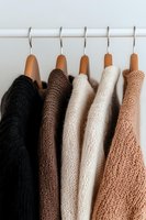 Cozy knitted sweaters in neutral earth tones hanging on wooden hangers, minimalist wardrobe organization showing texture and warm color palette.