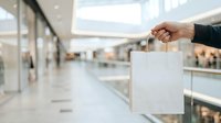 Person with medium skin tone holding blank white shopping bag in modern mall corridor with blurred retail storefronts, suitable for commerce and retail marketing campaigns.