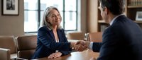 Senior businesswoman with gray hair shaking hands with male colleague in modern office meeting room during professional negotiation or partnership discussion.