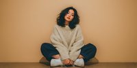 Young Asian woman with wavy hair sitting cross-legged against beige wall, wearing oversized knit sweater and blue jeans, conveying calm confidence.