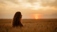 Young woman with long curly hair standing in golden wheat field during sunset, peaceful countryside landscape for wellness and freedom concepts.