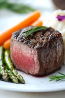Juicy medium rare filet mignon steak garnished with fresh rosemary, served with asparagus and baby carrots on white plate, close up view of gourmet dinner.