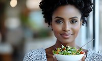 Young African American woman with natural curly hair smiling while holding fresh salad bowl in casual dining setting. Healthy eating and wellness lifestyle concept.