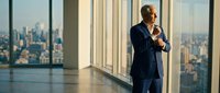 Professional businessman in navy suit standing by floor-to-ceiling windows overlooking city skyline, contemplating business decisions in modern office.