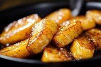 Caramelized banana slices sautéed in butter and brown sugar on black pan, close-up view showing golden crispy edges and glossy surface texture for food blogs.