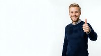 Young Caucasian man with blonde hair wearing dark navy sweater giving thumbs up gesture with confident smile against white background.