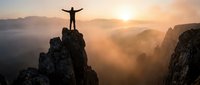 Silhouette of triumphant hiker with raised arms standing on rocky mountain peak at golden sunrise, celebrating success and freedom in dramatic landscape.