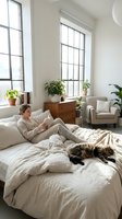 Woman in casual beige loungewear relaxing on bed with tabby cat in bright, minimalist bedroom featuring large windows, wooden furniture, potted plants and neutral decor.