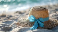 Elegant straw sun hat with turquoise ribbon bow resting on sandy beach with ocean waves in background for summer vacation and tropical travel concepts.