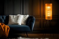 Moody interior with navy blue velvet sofa, orange throw blanket and decorative pillows against dark wall, illuminated by warm glowing floor lamp.