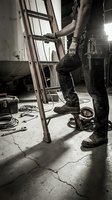 Construction worker climbing ladder at industrial worksite with tools and equipment scattered on concrete floor.