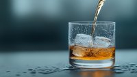 Amber liquid pouring into rocks glass with ice cubes, creating dynamic splash and bubbles against dark background, dramatic lighting highlights drink movement.