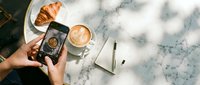Overhead view of person photographing croissant and latte coffee on marble table with smartphone, capturing food styling for social media content.