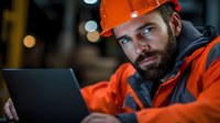 Bearded construction worker in orange safety helmet and high-visibility jacket using laptop computer at industrial site during evening shift operations.
