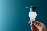 Hand holding light bulb with graduation cap against turquoise background, representing education, innovation and bright ideas in learning concept.