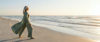 Young woman in flowing green dress walking along sandy beach at sunset with ocean waves and golden light creating peaceful coastal atmosphere for wellness content.
