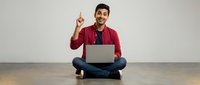 Young Hispanic man in red cardigan sitting cross-legged with laptop pointing finger up having eureka moment against neutral background for business concepts.
