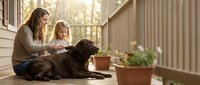 Woman and young girl petting brown dog on porch with potted plants, outdoor family bonding moment with pet.