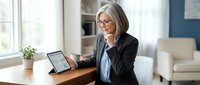 Mature businesswoman with gray hair and glasses working on tablet at home office desk, professional remote work setup with modern interior.