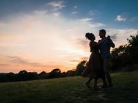 Silhouetted couple dancing in open field at sunset, man and woman embracing outdoors during golden hour with dramatic sky and trees, romantic moment for wedding and engagement content.