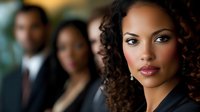 Young African American businesswoman with curly hair and professional makeup in corporate office setting, colleagues in background, confident expression.
