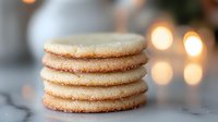 Stack of homemade sugar cookies with sparkly crystallized edges on marble surface against soft glowing bokeh lights creating cozy holiday atmosphere.