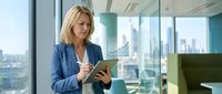 Professional blonde businesswoman using digital tablet in modern office with city skyline view for corporate technology and workplace productivity.