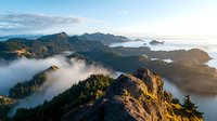 Majestic mountain peaks emerge through low-lying clouds at sunrise, creating dramatic landscape with golden light illuminating rocky cliffs and evergreen forest slopes.