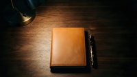 Closed tan leather notebook with black pen on dark wooden desk surface, illuminated by warm overhead lighting for business and professional use.