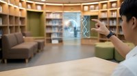 Young male professional with East Asian appearance demonstrating augmented reality technology on digital interface in modern library setting with wooden shelving and contemporary furniture.