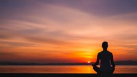 Man silhouette meditating in lotus pose at sunset by lake with dramatic orange purple sky reflecting on calm water surface for wellness mindfulness concepts.