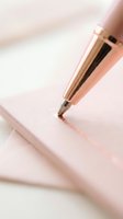 Rose gold pen writing on pink notebook, close-up detail of ink tip touching paper surface for writing, journaling, note-taking, and stationery design projects.