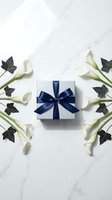 Elegant silver gift box with navy blue satin bow surrounded by white calla lilies and ivy on marble background, luxury present concept.