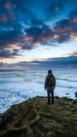 Male hiker in dark jacket standing on grassy cliff overlooking turbulent ocean waves at dramatic sunset with blue and orange clouds. Adventure and travel concept.