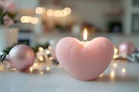 Pink heart-shaped candle glowing with warm light, surrounded by rose gold Christmas ornaments and bokeh lights on white surface, creating romantic atmosphere.
