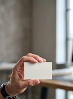 Caucasian male hand holding blank white business card in modern office environment with natural window light for corporate branding and professional networking purposes.