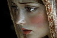 Young woman with freckles and natural makeup wearing traditional lace veil, side profile portrait showing delicate features and rosy cheeks against dark background.