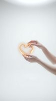 Hands gently holding glowing golden heart symbol against soft white background representing love, care, compassion and emotional wellness concepts.
