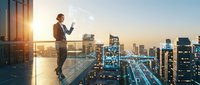 Young businessman using smartphone on modern office balcony overlooking illuminated city skyline at sunset for corporate communication and urban lifestyle.