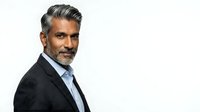 Mature man with salt-and-pepper hair and beard, South Asian ethnicity, wearing dark blazer and light blue shirt, professional business portrait against white background.