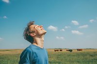 Young Caucasian man in blue t-shirt laughing with head tilted back against blue sky and prairie landscape with grazing cattle, expressing pure joy and freedom.