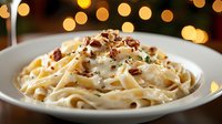 Creamy fettuccine pasta with chopped pecans and fresh herbs garnish served on white plate against warm bokeh lights background, restaurant setting.