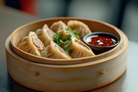 Steamed dumplings in traditional bamboo basket served with spicy chili sauce and fresh herbs, close up view of Asian cuisine appetizer.