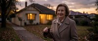 Smiling senior Caucasian woman in beige coat standing outside suburban home at sunset with warm glowing windows creating cozy residential atmosphere.