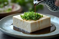 Fresh tofu block with green onions being drizzled with soy sauce on ceramic plate, traditional Japanese cuisine preparation for healthy meal.