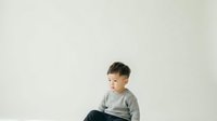 Young Asian boy, approximately 3-5 years old, wearing gray long-sleeve shirt and dark pants, sitting against white background, looking to side with neutral expression.