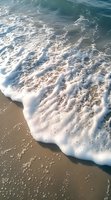 Gentle ocean waves with white foam washing up on sandy beach shore, creating natural patterns as water meets sand in soft morning sunlight.